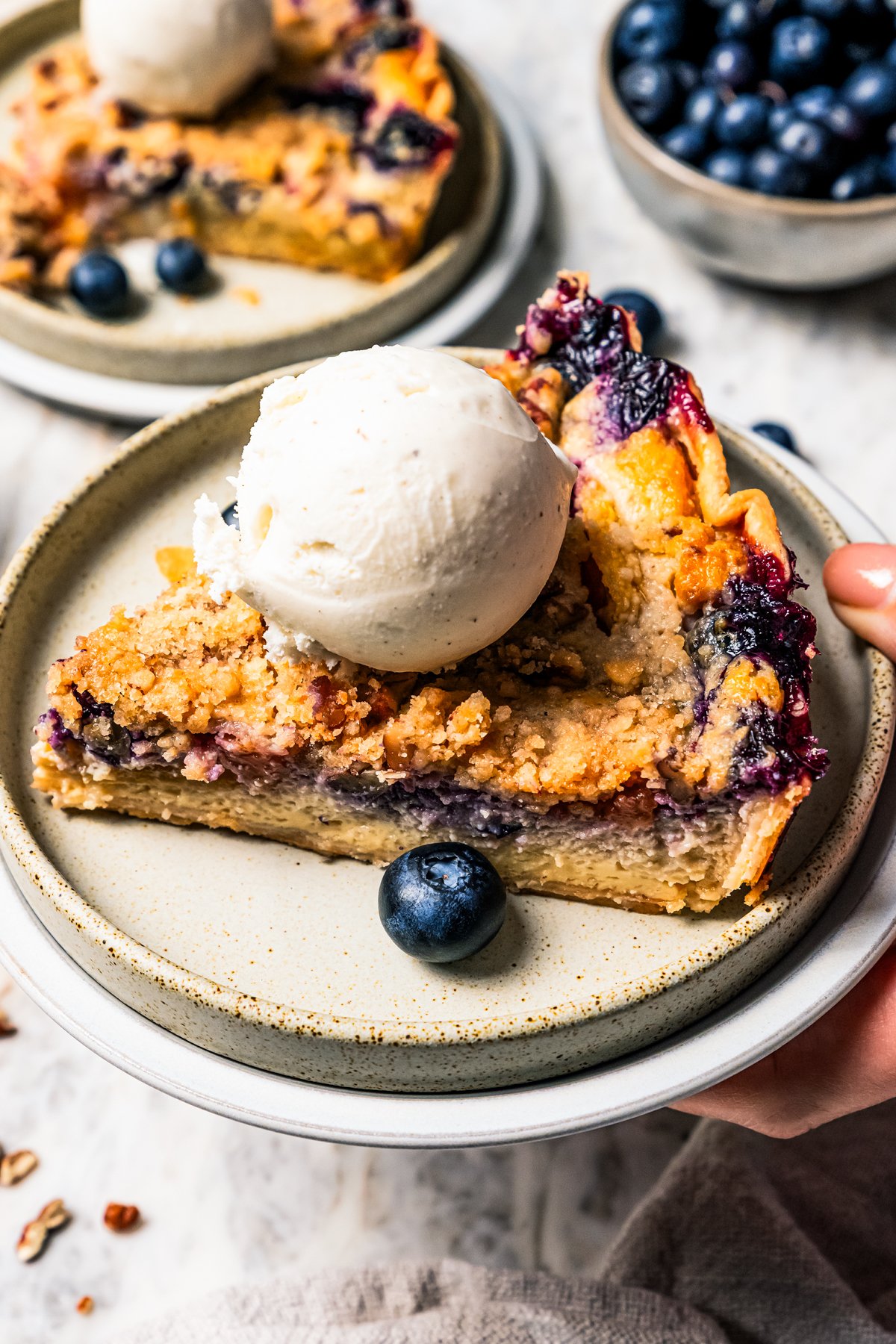 This peach blueberry pie is a fruit-filled custard pie bursting with juicy fresh peaches and blueberries, topped with a nutty pecan streusel. A slice of blueberry pie topped with a scoop of vanilla ice cream on a plate.