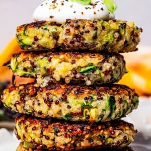 Five quinoa and zucchini fritters stacked on a plate and topped with a dollop of plain yogurt.