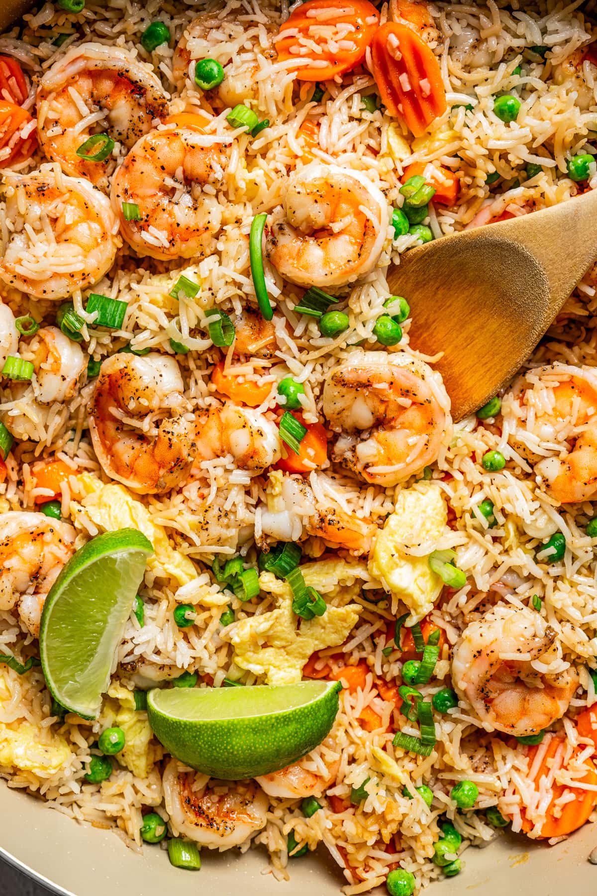 Close-up photo of shrimp fried rice in a skillet with a wooden spoon stirring through it. Two lime wedges are placed on top of the rice.