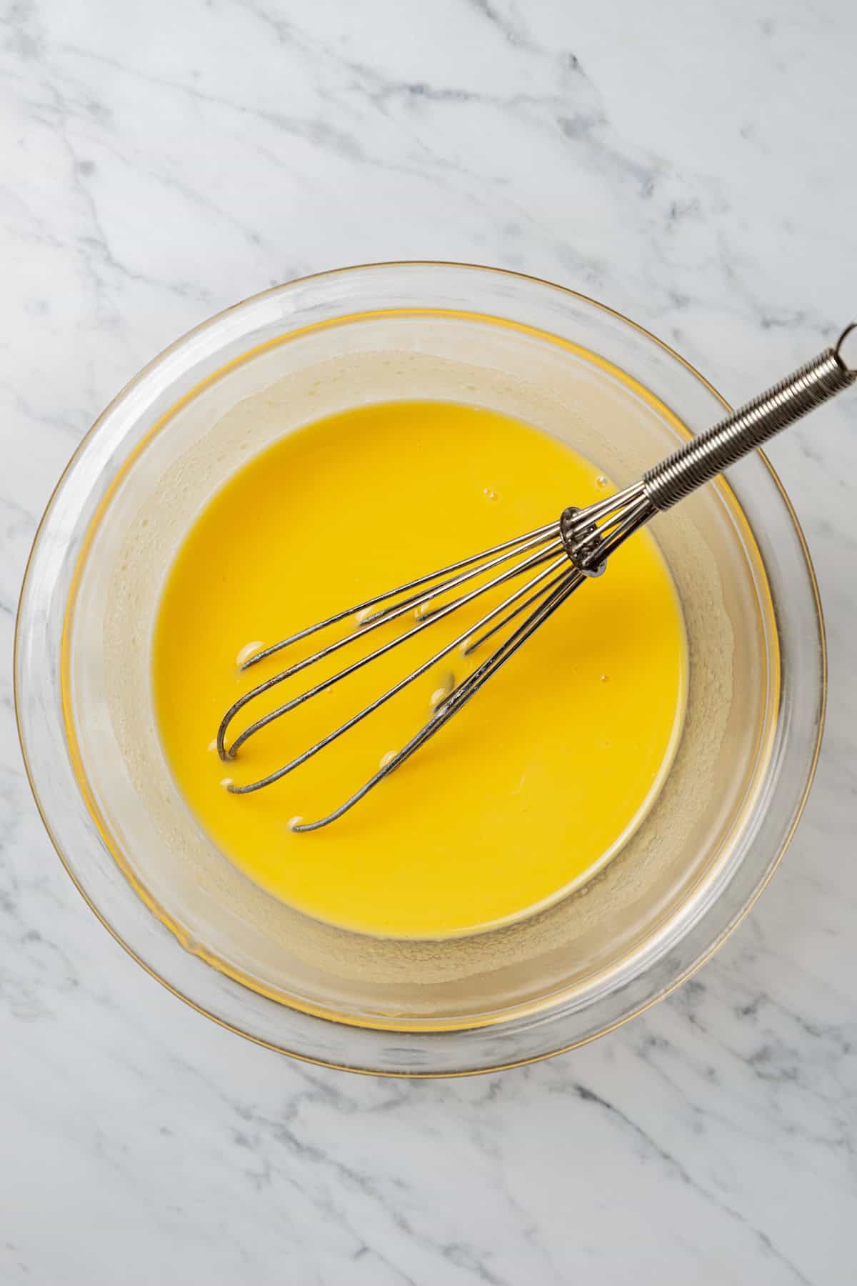 A whisk resting in a bowl of beaten eggs.