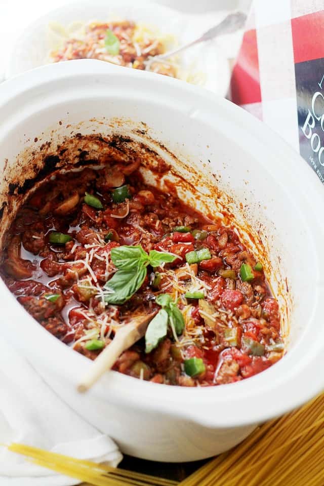 Side view of a slow cooker insert filled with spaghetti meat sauce and garnished with fresh basil leaves.