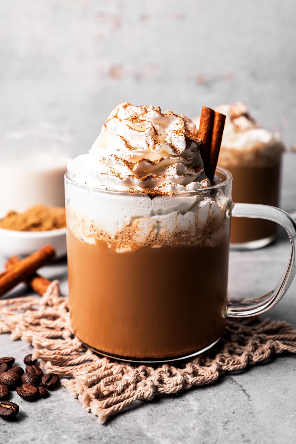 A cinnamon dolce latte in a glass mug garnished with whipped cream and a cinnamon stick, dusted with cinnamon, with a second latte in the background.