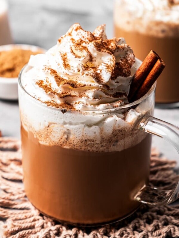 A cinnamon dolce latte in a glass mug garnished with whipped cream and a cinnamon stick, dusted with cinnamon, with a second latte in the background.
