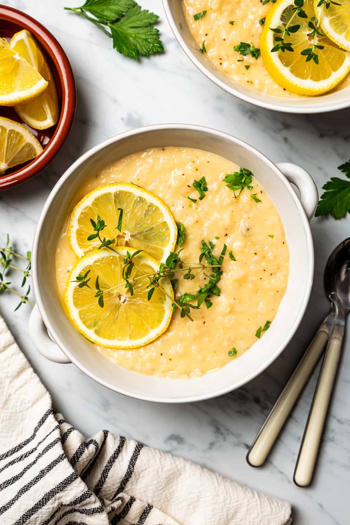 A bowl of lemon rice soup garnished with lemon slices and thyme sprigs is next to a small dish filled with lemon wedges and a second bowl of soup.