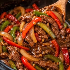 Beef stir fry with bell peppers and onions being scooped from the slow cooker with a wooden spoon.