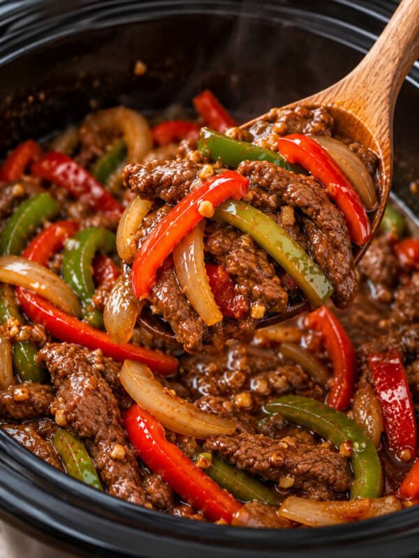 Beef stir fry with bell peppers and onions being scooped from the slow cooker with a wooden spoon.