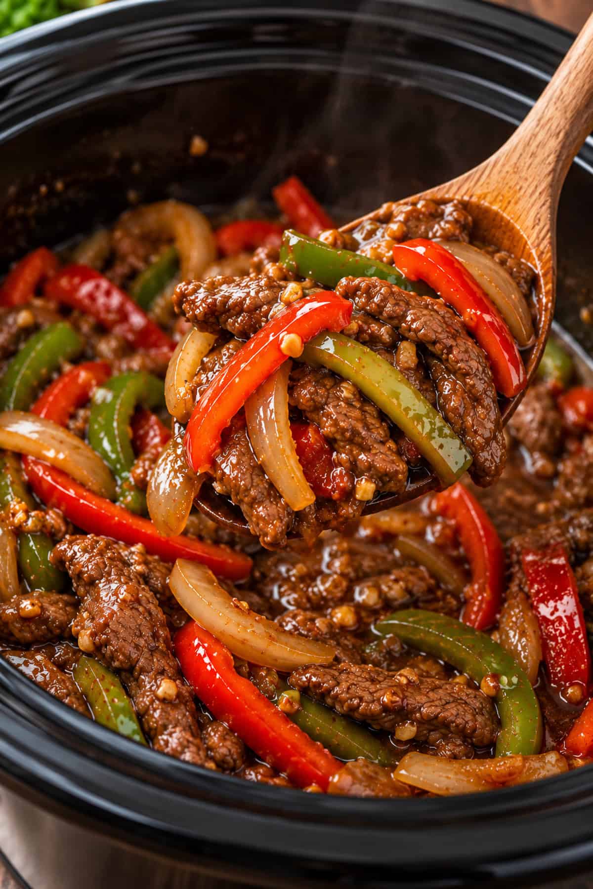 Beef stir fry with bell peppers and onions being scooped from the slow cooker with a wooden spoon.