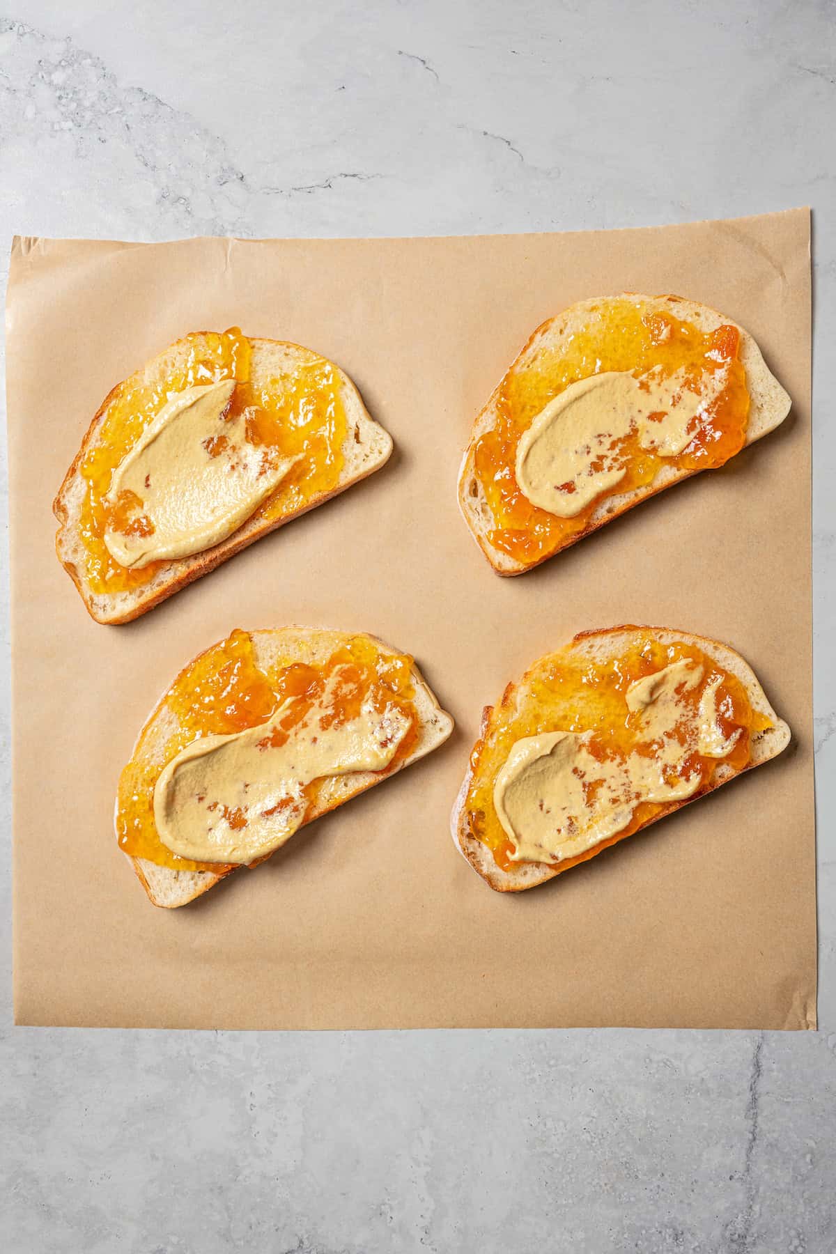 Overhead view of bread slices spread with apricot jam and Dijon mustard on a piece of parchment paper.