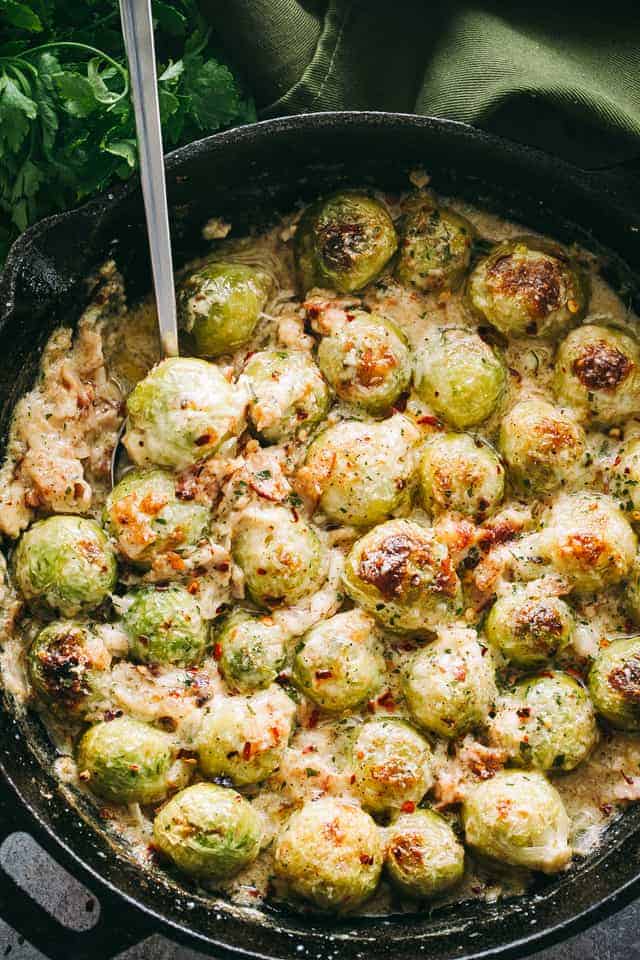 A skillet of cheesy Brussels sprouts with bacon and cheese sauce, with a spoon for serving.