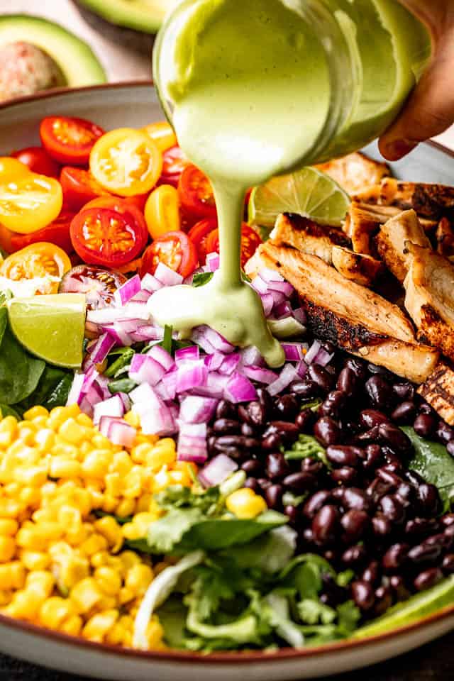 Pouring avocado dressing over a salad with chicken, corn, black beans, salad greens, red onions, and tomatoes.