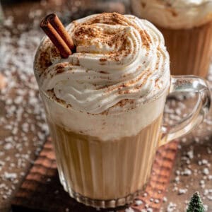 A Starbucks eggnog latte garnished with whipped cream, ground nutmeg, and a cinnamon stick in a glass mug, with second latte in the background.