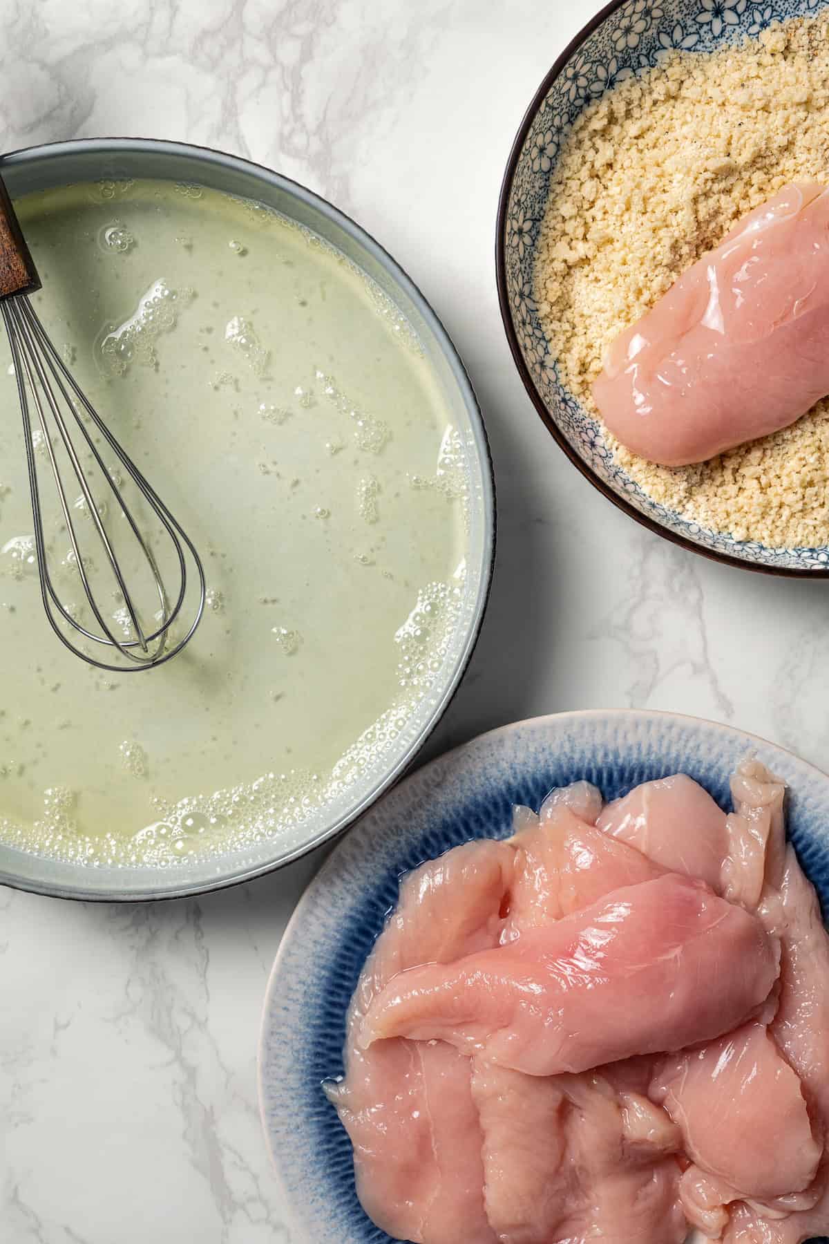 Overhead view of a breading station with chicken cutlets on a plate next to a bowl of beaten egg whites, and piece of chicken in a bowl with breadcrumbs.