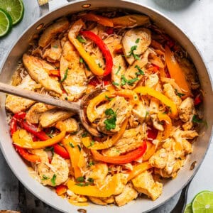 An overhead view shows a wooden spoon resting in a skillet of cooked sliced chicken, bell peppers, and onions.