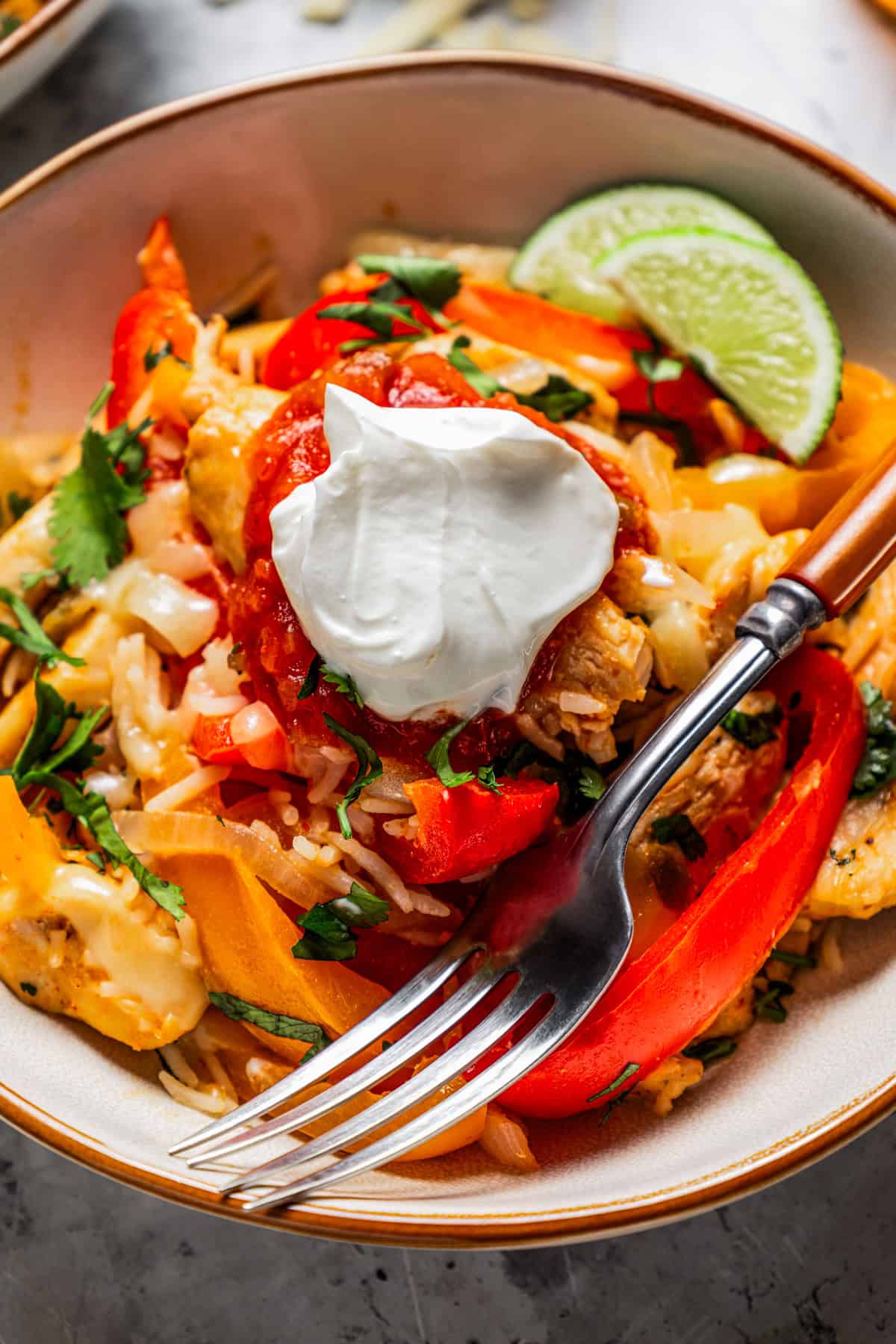 Close up of a chicken fajita bowl garnished with lime wedges and a dollop of sour cream, with a fork.