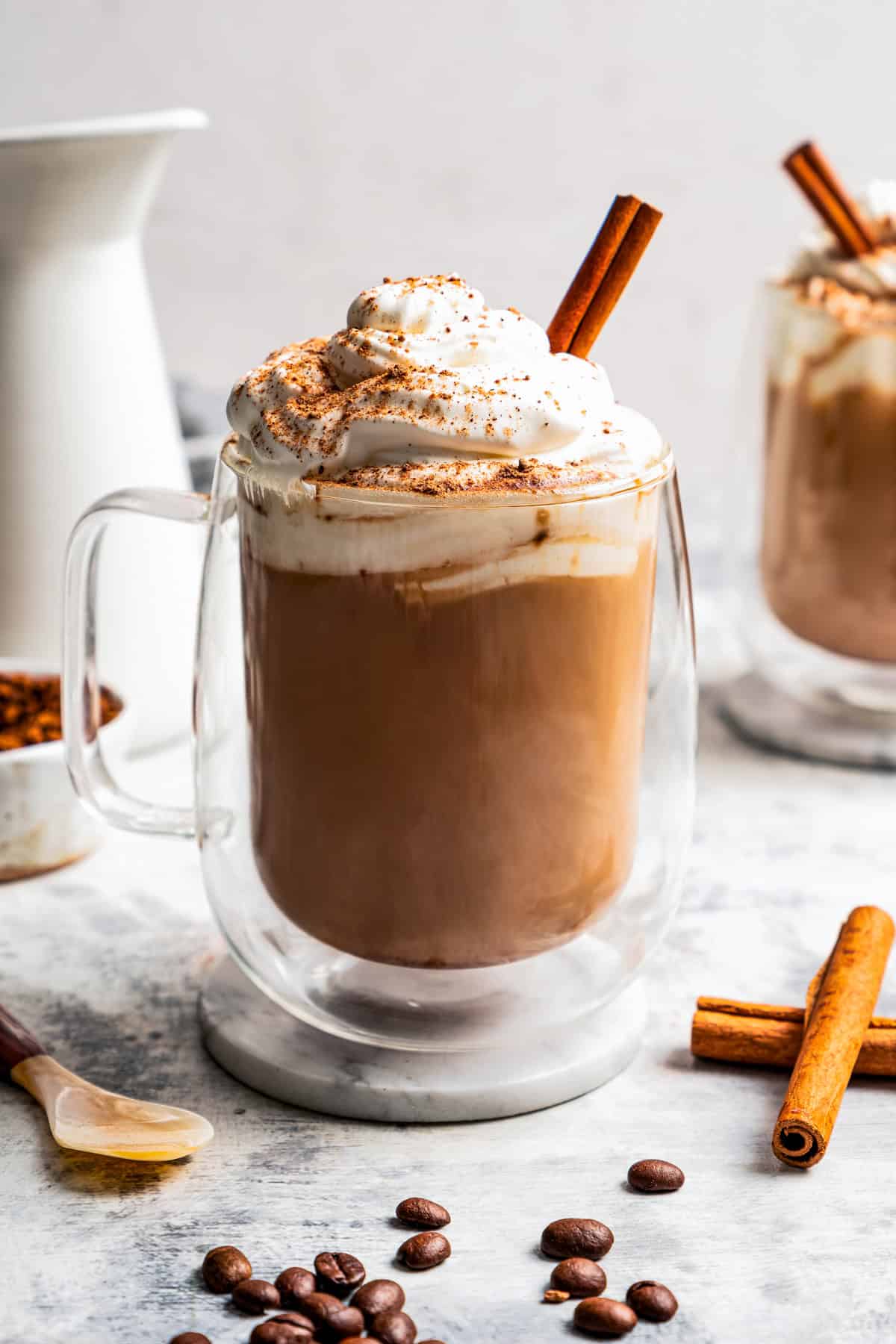 A Starbucks eggnog latte garnished with whipped cream, ground nutmeg, and a cinnamon stick in a glass mug, with a second latte in the background.