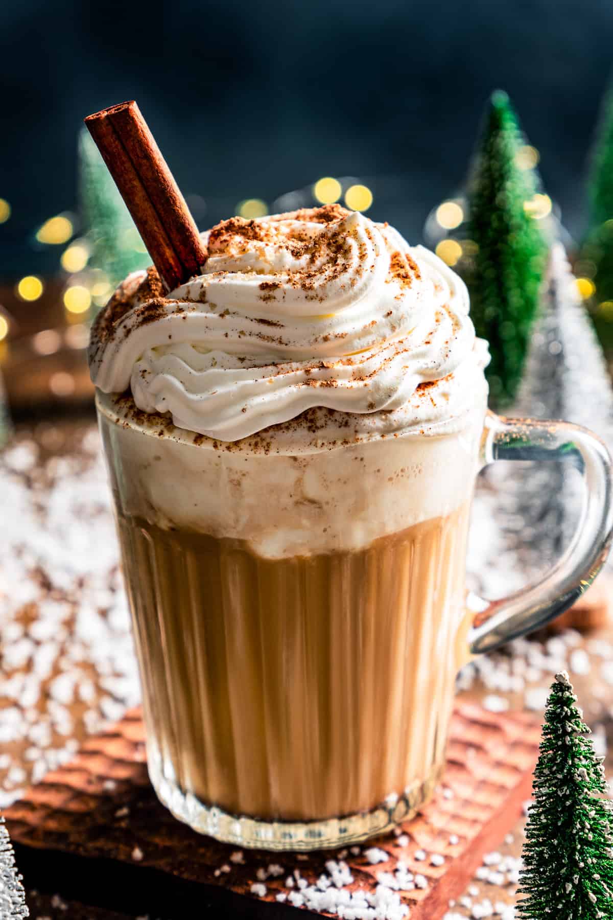 A Starbucks eggnog latte garnished with whipped cream, ground nutmeg, and a cinnamon stick in a glass mug, with a festive holiday Christmas tree backdrop.