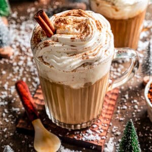 A Starbucks eggnog latte garnished with whipped cream, ground nutmeg, and a cinnamon stick in a glass mug, with second mug of latte in the back.