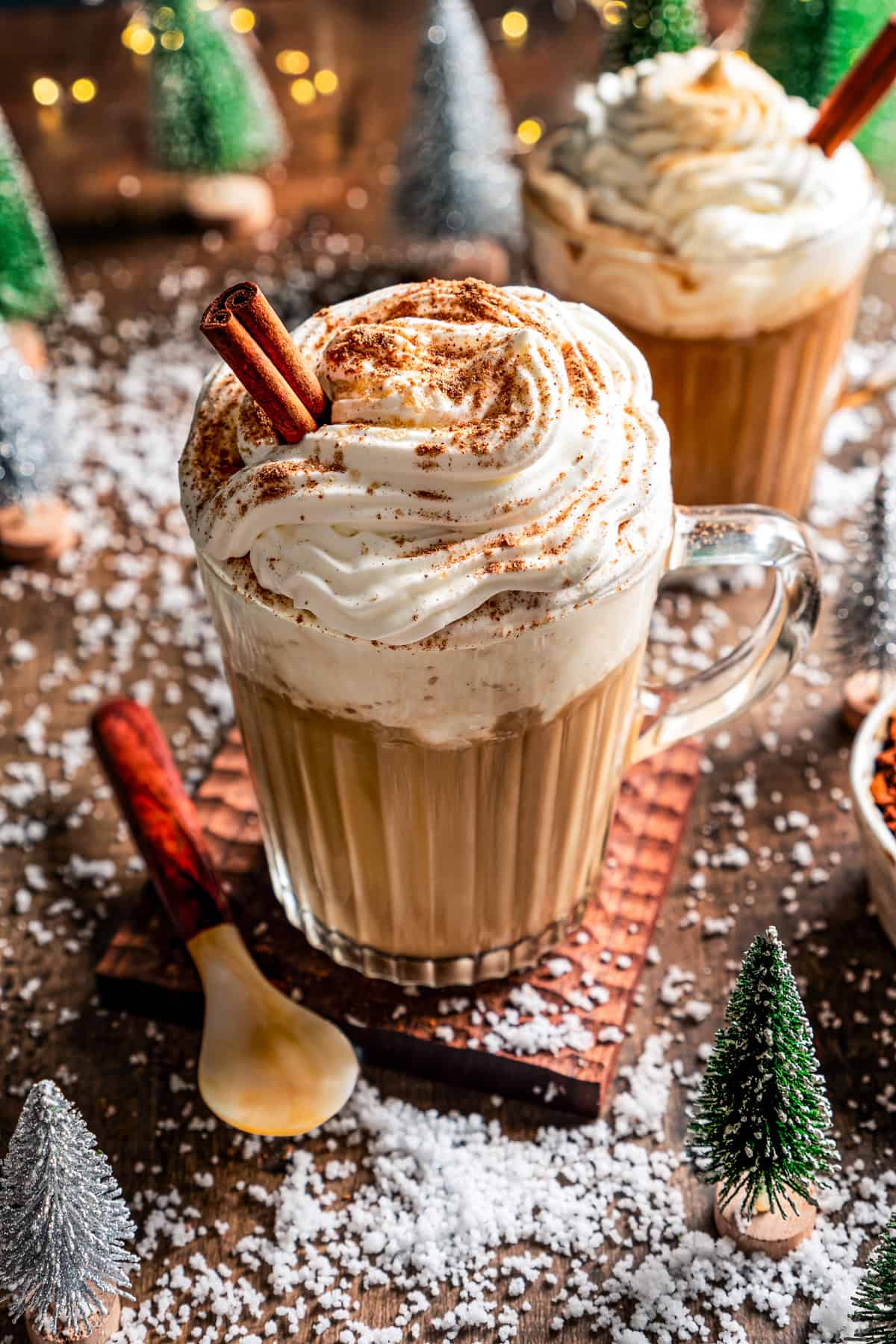 A Starbucks eggnog latte garnished with whipped cream, ground nutmeg, and a cinnamon stick in a glass mug, with second mug of latte in the back.