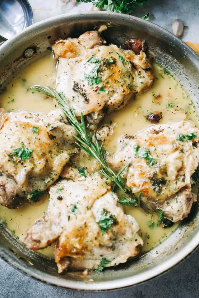 Overhead shot of four chicken thighs in garlic sauce, with a garnish of a rosemary sprig and chopped parsley.