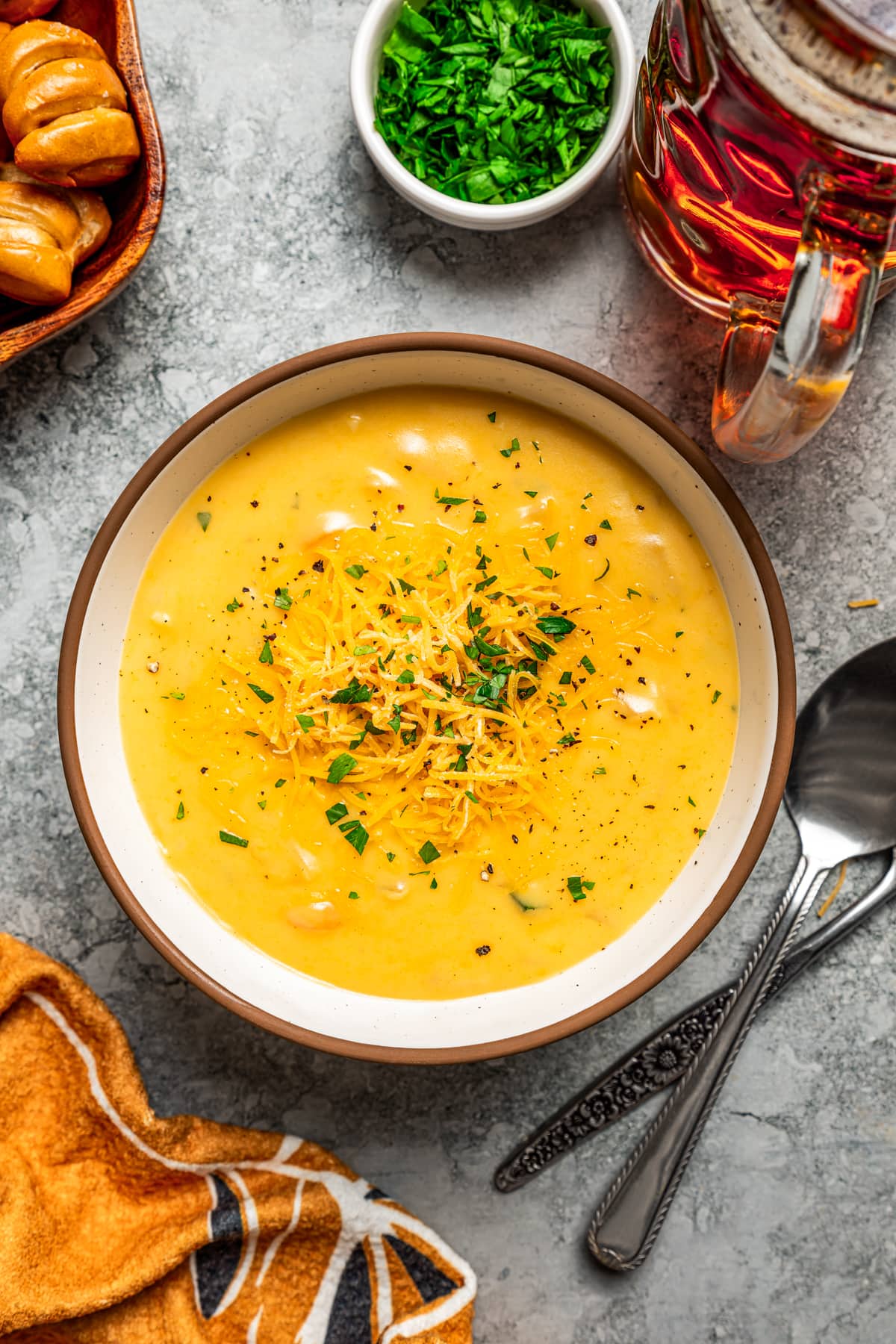 Overhead view of a bowl of beer cheese soup with a spoon on a countertop surrounded by ingredients.
