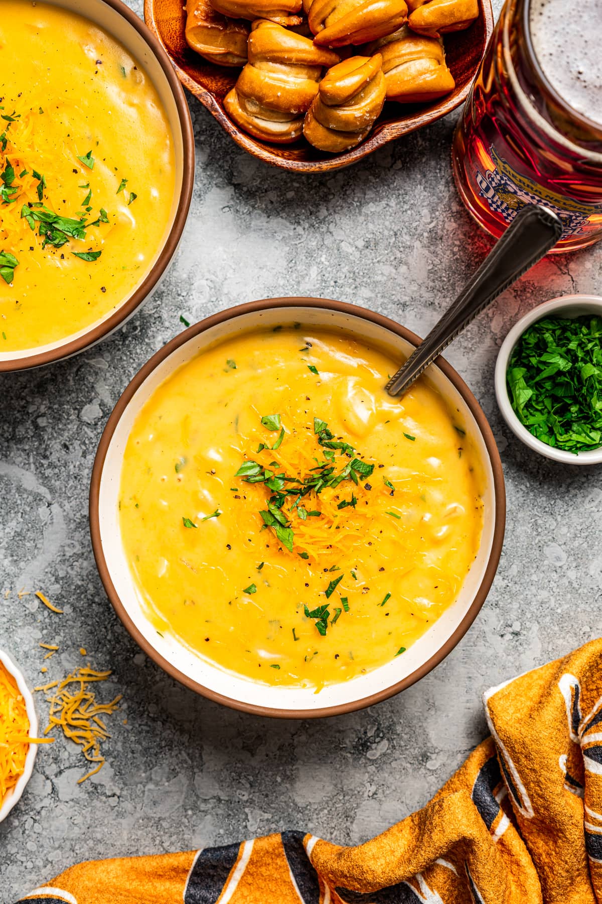 Overhead image of a bowl of beer cheese soup with a spoon on a countertop surrounded by ingredients.
