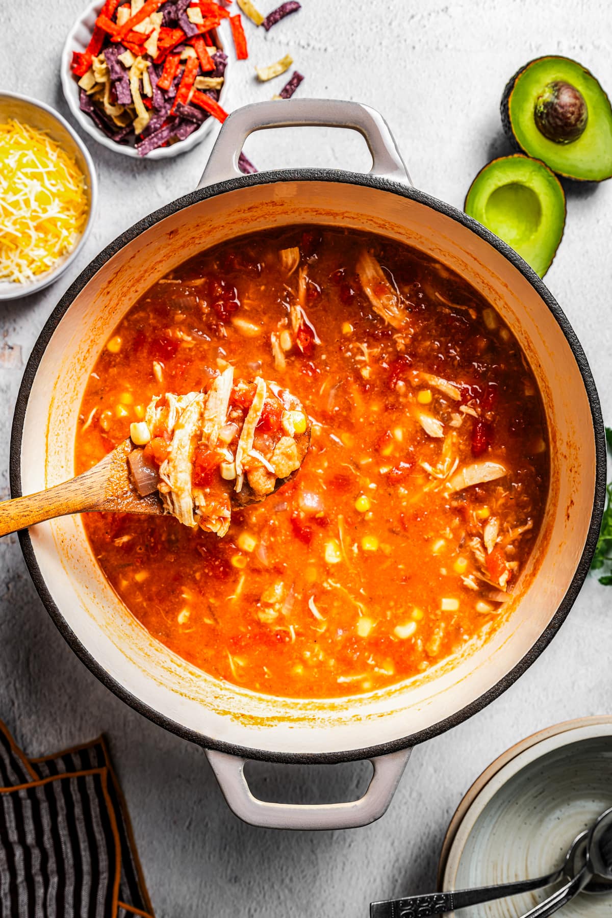 Chicken tortilla soup in a pot near tortilla strips, cheese, and avocado.