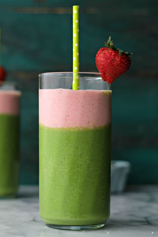 A glass filled with a layered Green Berry Smoothie with a green straw and strawberry on the rim