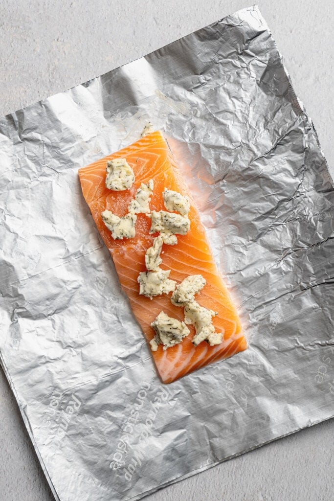 A salmon filet placed over a foil sheet, topped with lemon pepper butter.