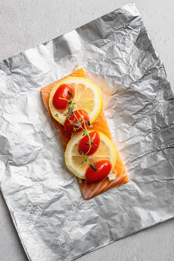A salmon filet placed over a foil sheet, topped with lemon pepper butter, lemon slices, grape tomatoes, and a thyme sprig.