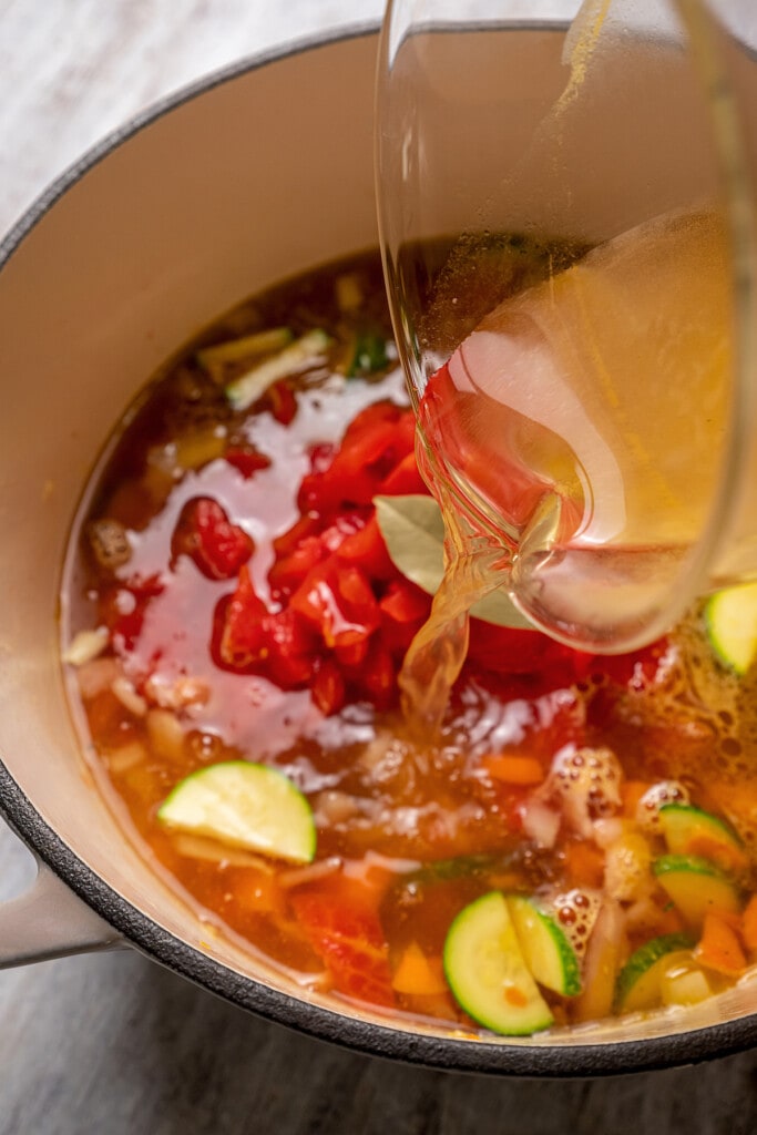 Vegetable broth poured over minestrone soup ingredients in a large pot.