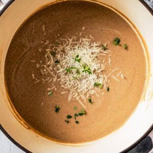 Overhead view of creamy mushroom soup in a large pot garnished with shredded parmesan and fresh herbs.