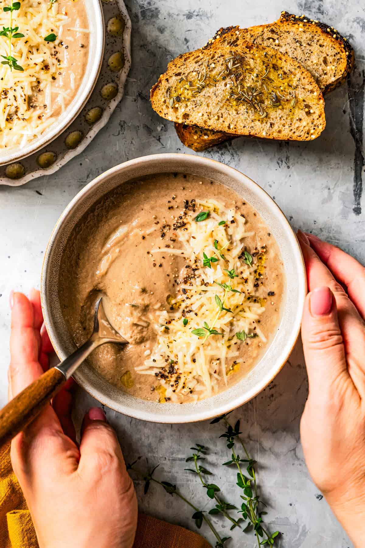 Overhead view of two hands touching a bowl of creamy mushroom soup garnished with shredded parmesan and fresh herbs, next to two bread slices.