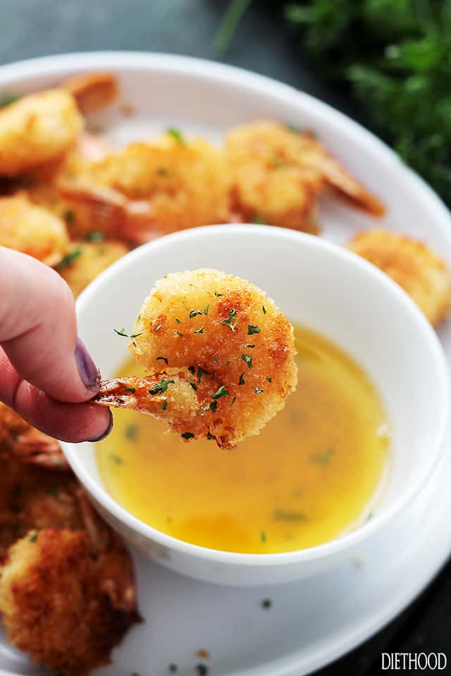 Hand holding a baked battered shrimp over a bowl of garlic sauce.
