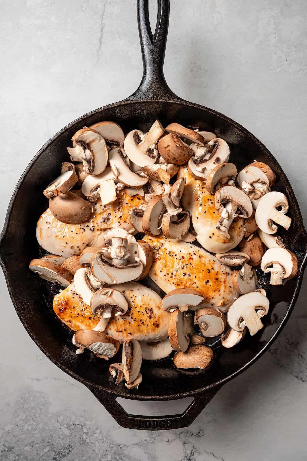 Sliced mushrooms added to a skillet with seared chicken breasts.