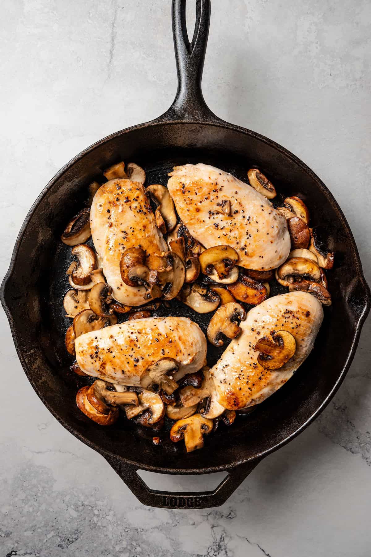 Overhead view of four browned chicken breasts surrounded by sautéed mushrooms in a skillet.