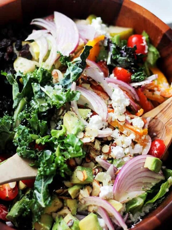 A wood bowl filled with kale, onions, feta, tomatoes, avocados, almonds and more