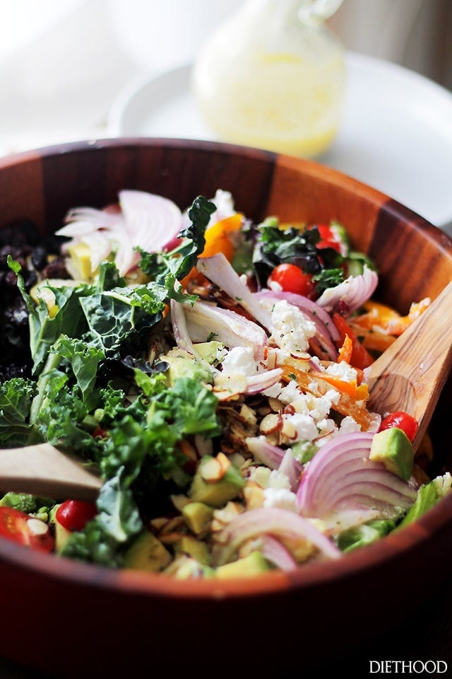 A wood bowl filled with kale, onions, feta, tomatoes, avocados, almonds and more