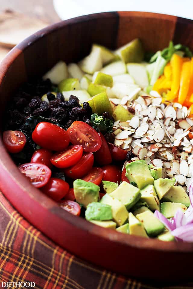 A wooden bowl with the ingredients for a hearty kale salad including tomatoes, avocado, and almonds