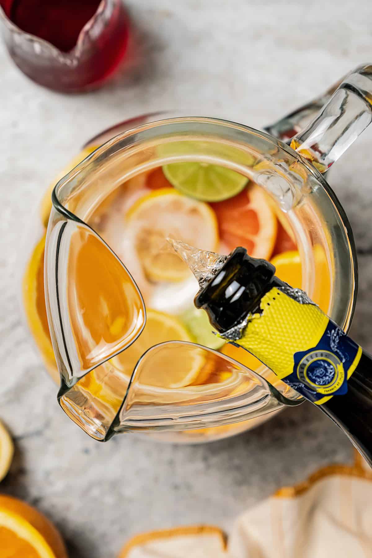 Overhead view of prosecco being poured from the bottle into a pitcher filled with sliced citrus.