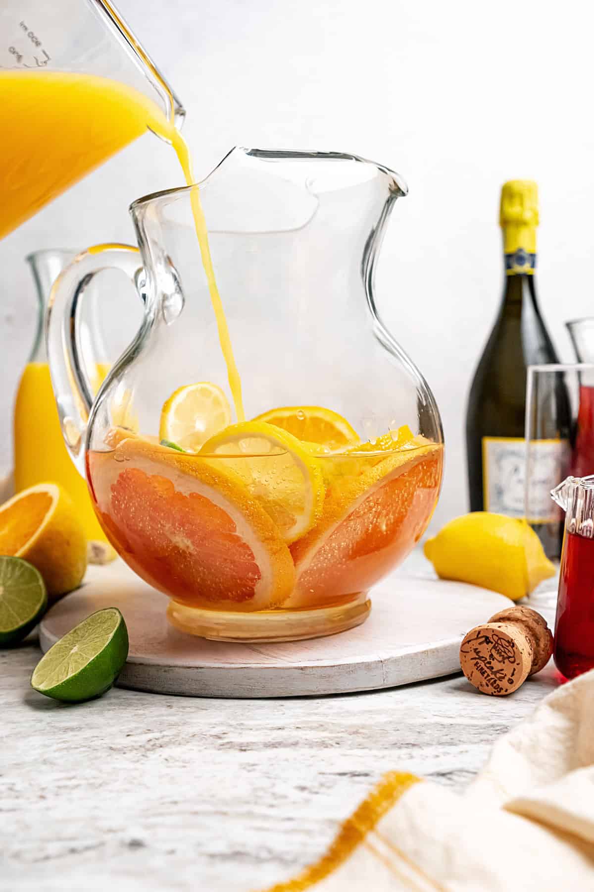 Orange juice being poured into a pitcher containing citrus slices and prosecco.