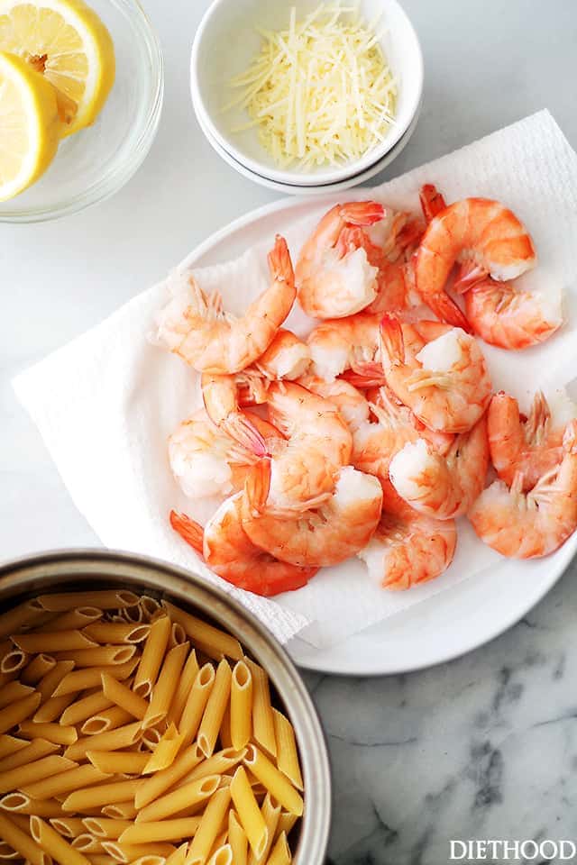 Overhead view of a plate of shrimp next to a bowl of penne pasta, a smaller bowl of parmesan, and lemon slices.