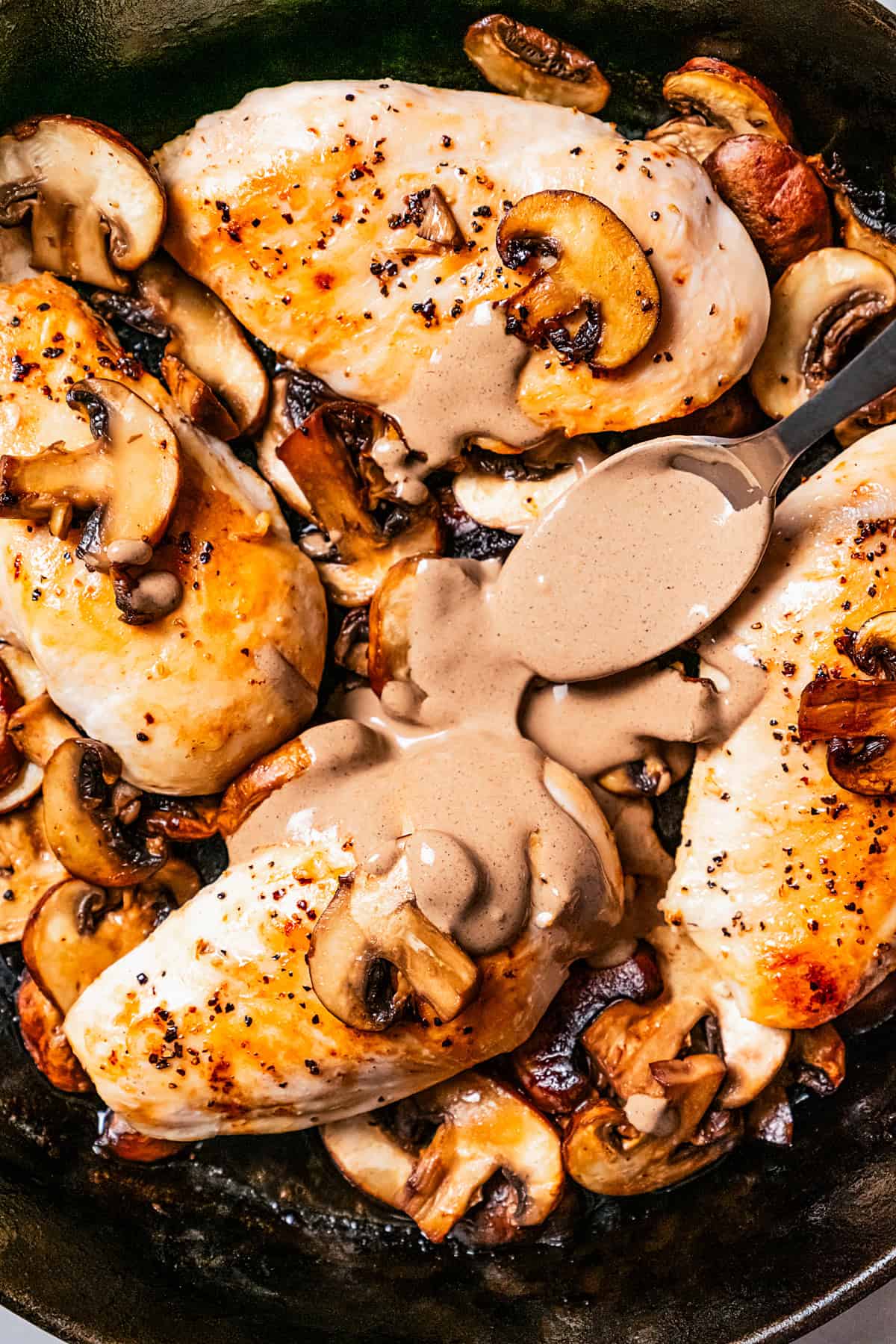 A spoon drizzling Dijon mushroom sauce over chicken Dijon in a cast iron skillet.