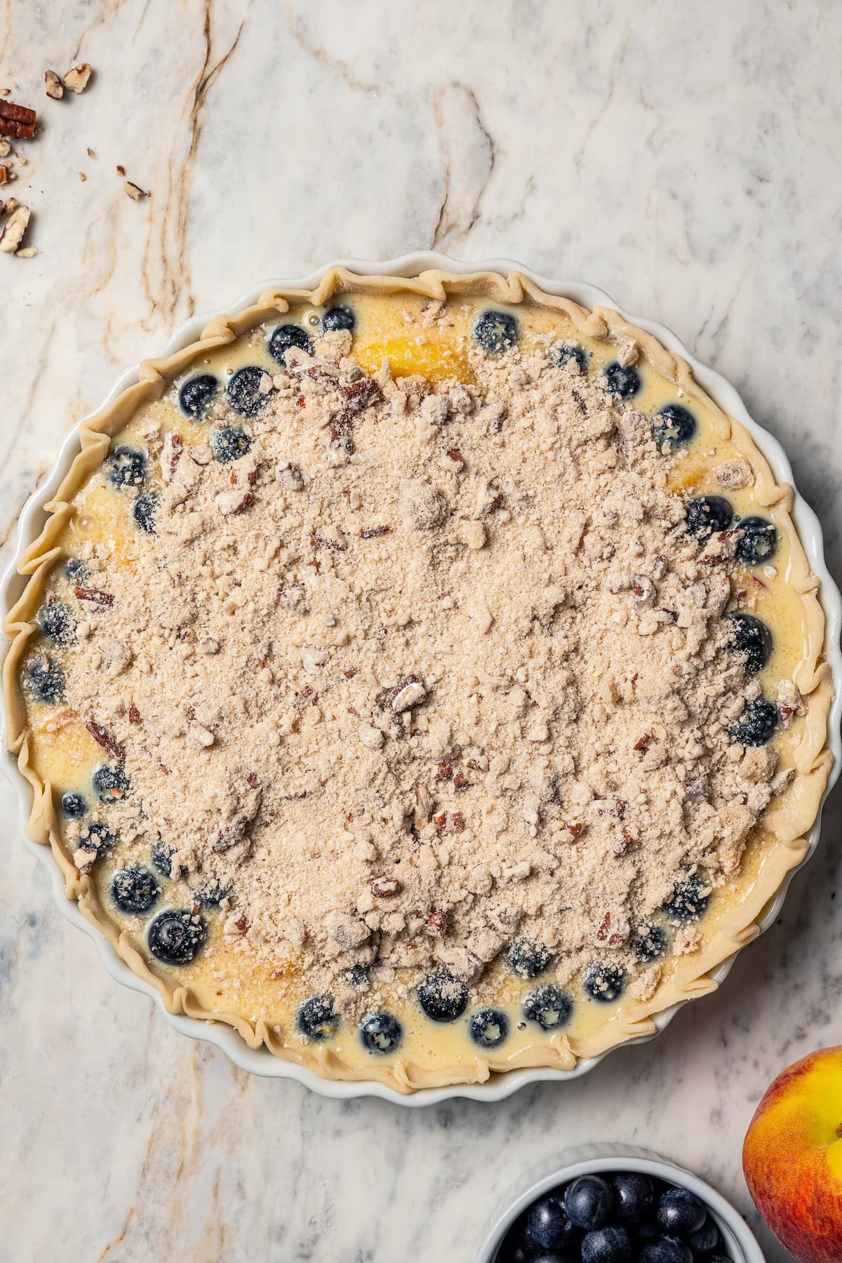 This peach blueberry pie is a fruit-filled custard pie bursting with juicy fresh peaches and blueberries, topped with a nutty pecan streusel. Overhead view of an unbaked peach blueberry pie topped with streusel.