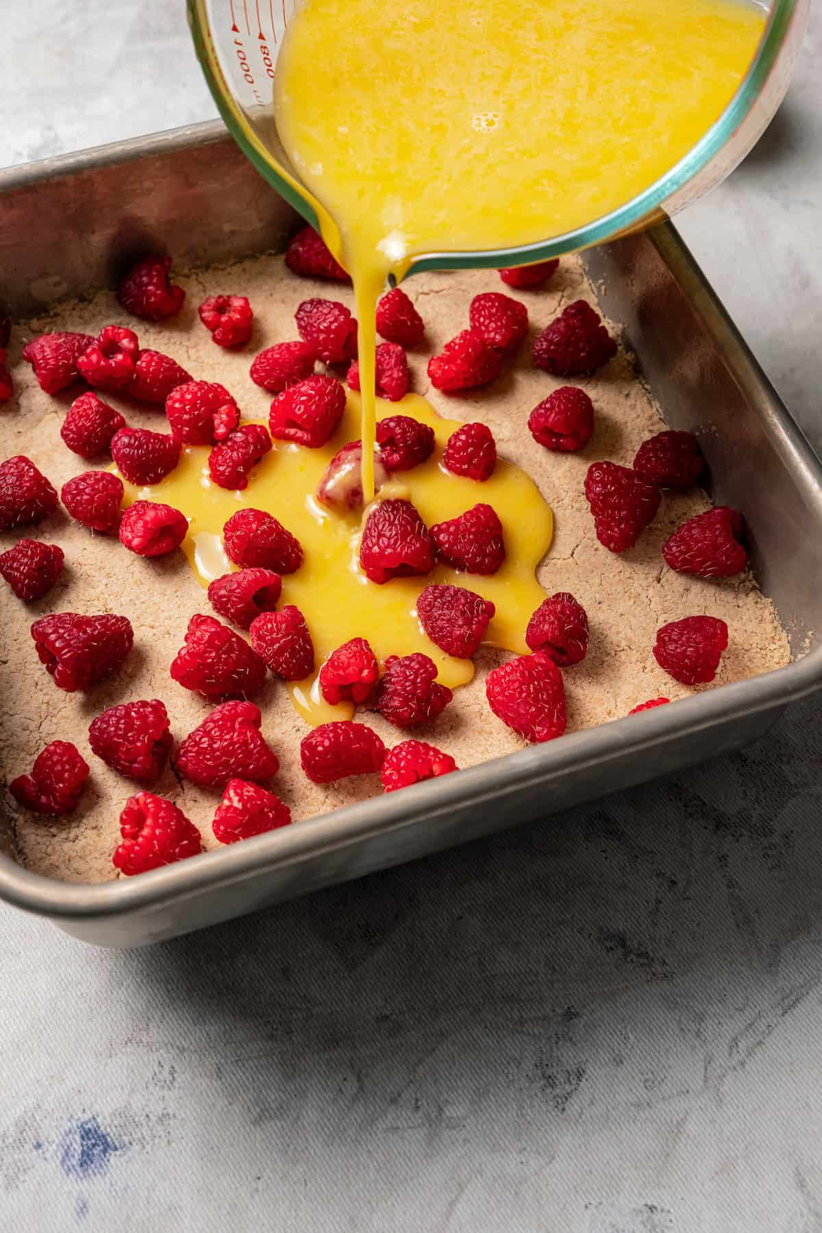 Pouring lemon bar filling over fresh strawberries on top of a cookie crust in a baking pan.