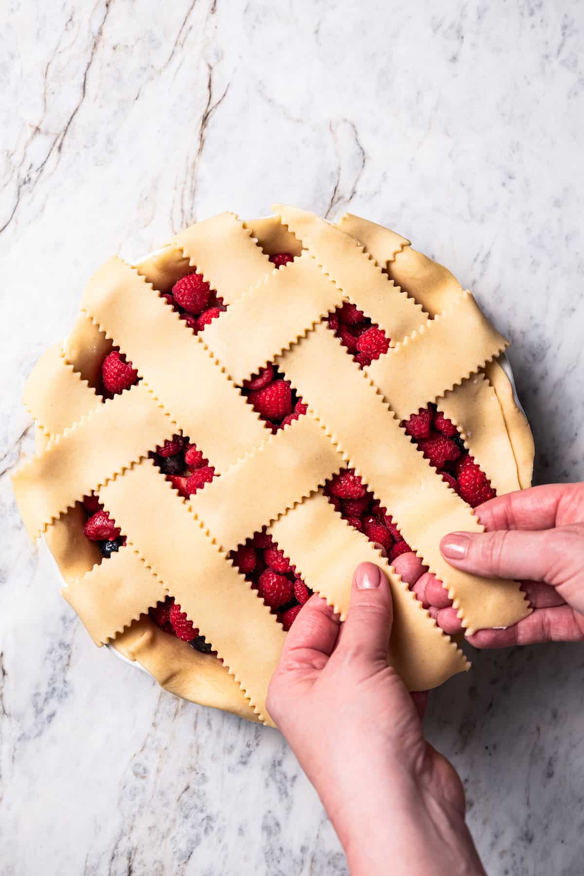 This easy mixed berry pie recipe combines three types of juicy berries with tender apples inside a light and flaky homemade pie crust. Two hands making a lattice pie.