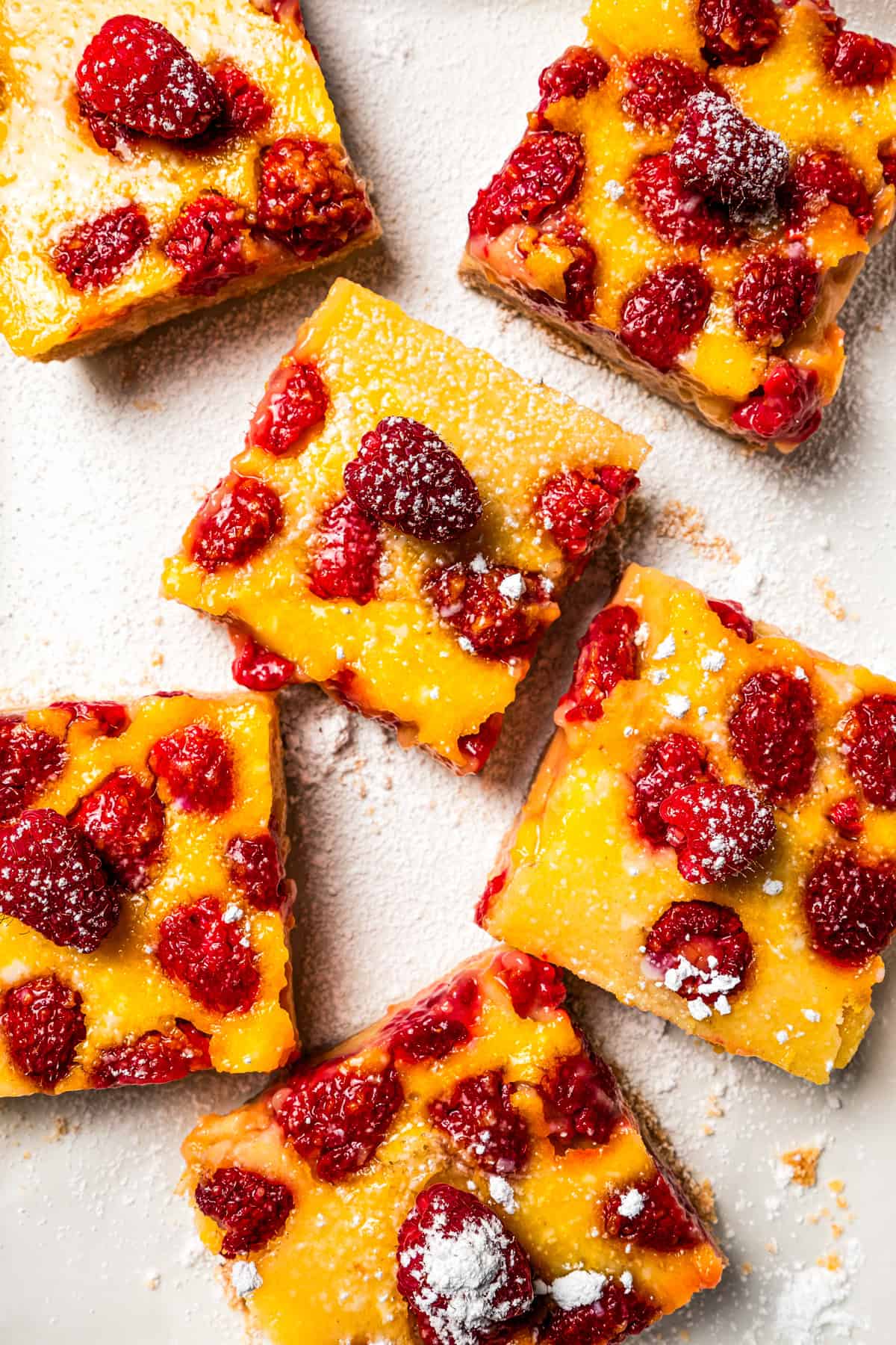 Lemon bars topped with raspberries and arranged on a white backdrop.