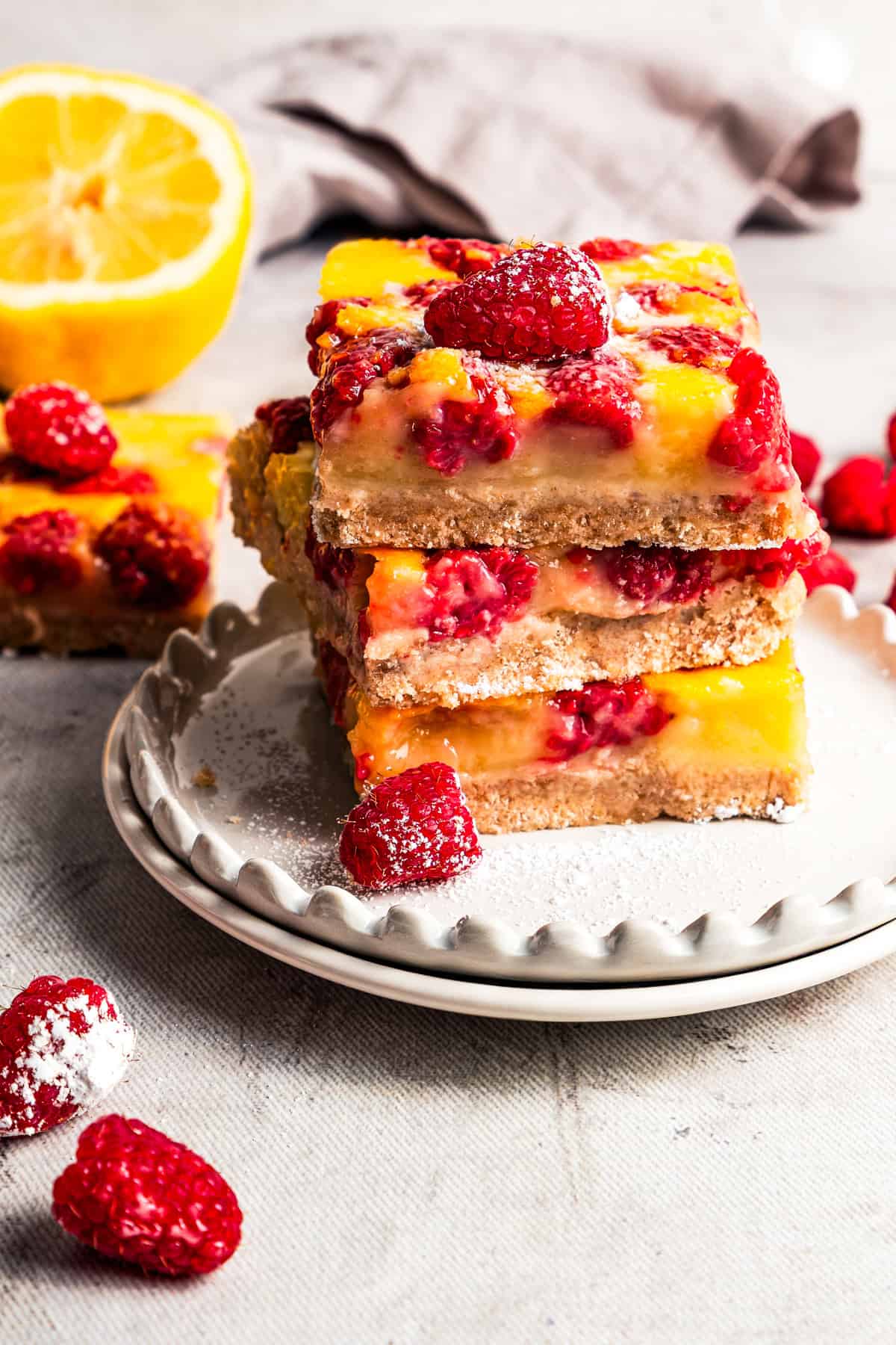 Three raspberry lemon bars stacked on a plate, dusted with powdered sugar, with fresh lemons and raspberries in the background. Photos and recipe by diethood.com