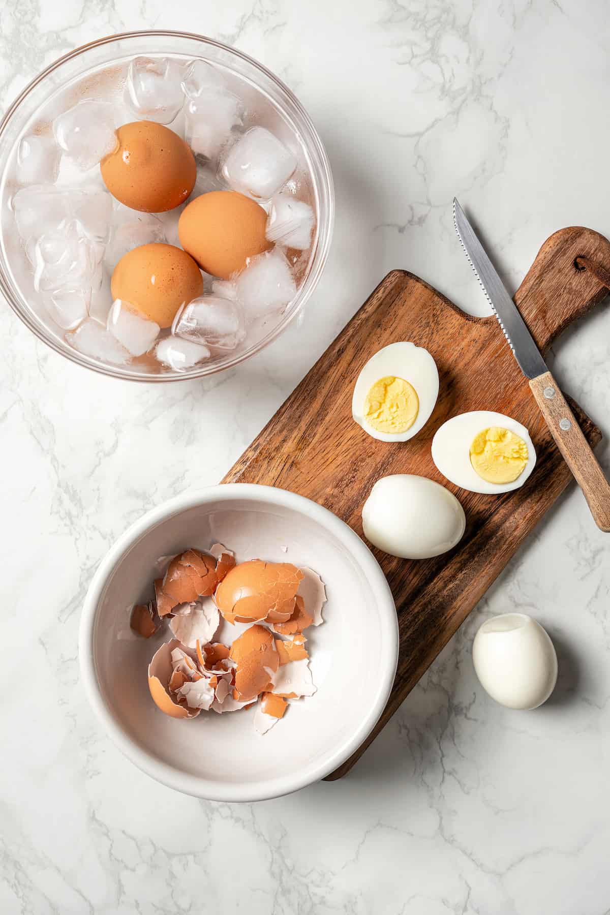 A bowl of eggs in ice water next to peeled hard boiled eggs on a wooden cutting board and a small bowl of shells.