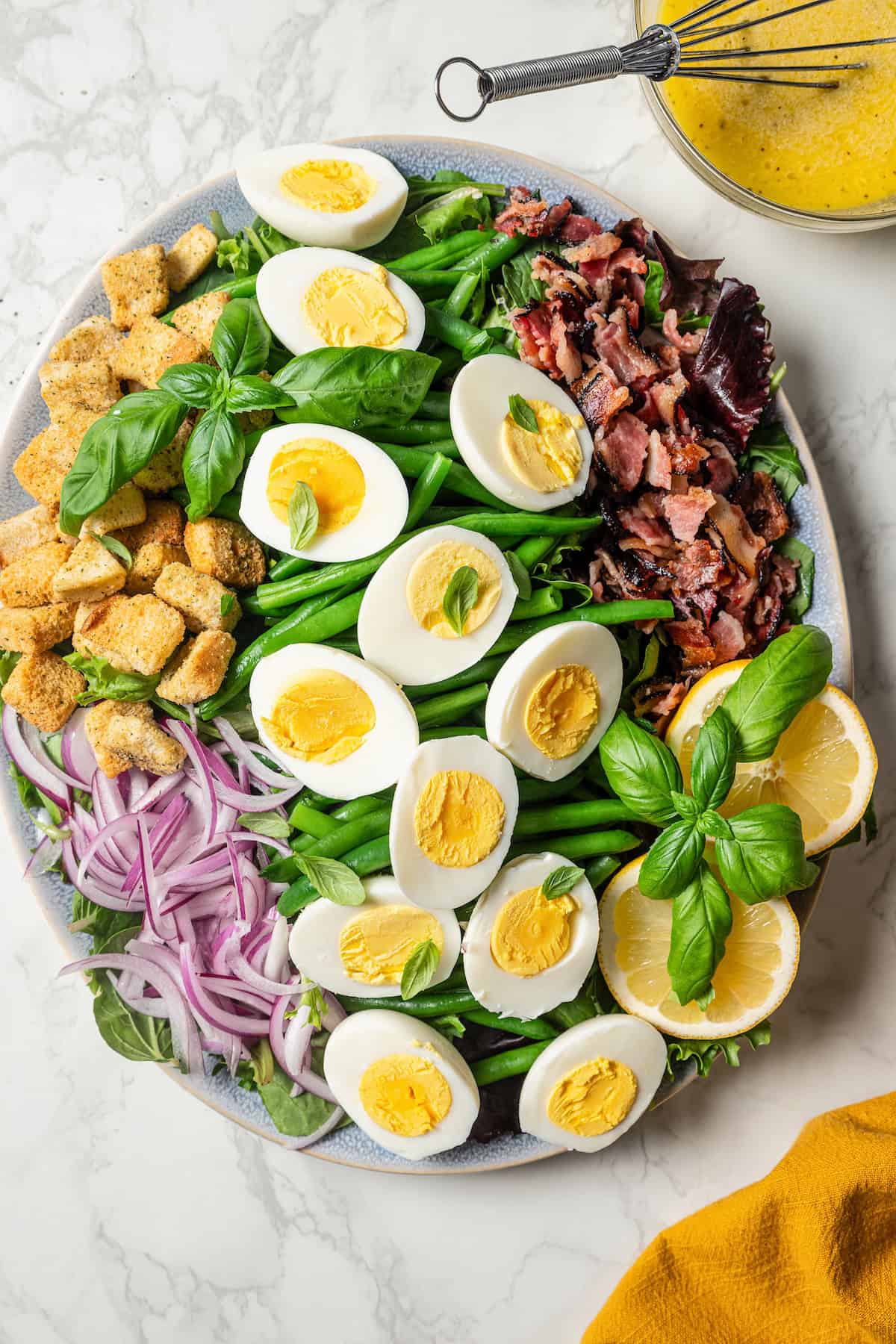 Overhead view of a green bean salad on a large platter topped with sliced hard boiled eggs, bacon, and croutons.