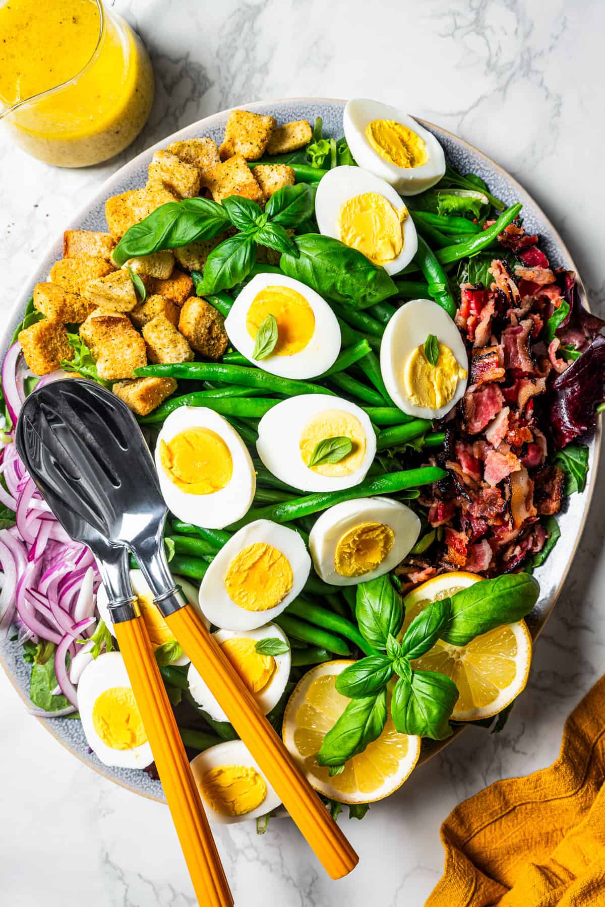 Overhead view of a green bean salad on a large platter topped with sliced hard boiled eggs, bacon, and croutons, with a set of salad tongs.